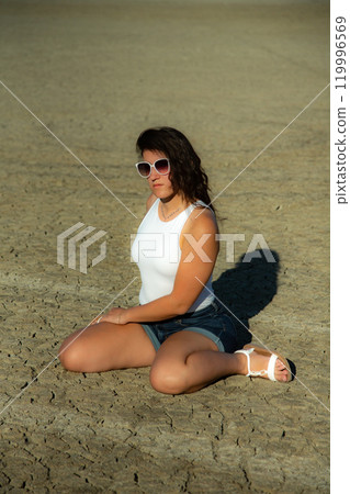 Vertical composition. Portrait young woman in sunglasses sits on dry desert ground 119996569