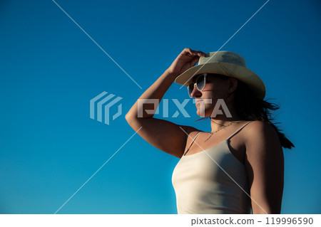 Portrait Young woman in white dress and sunglasses with hat posing against the backdrop of sky. Close-up Portrait Young woman in white dress and sunglasses with hat posing against the backdrop of sky. Close-up 119996590