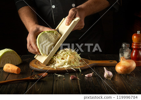 With careful precision, a chef shreds fresh cabbage on a wooden cutting board, surrounded by garlic, spices, and vibrant vegetables, creating the perfect ingredients for a delicious dish With careful precision, a chef shreds fresh cabbage on a wooden cutting board, surrounded by garlic, spices, and vibrant vegetables, creating the perfect ingredients for a delicious dish 119996784