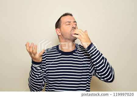 Bearded hispanic man in his 40s wearing a striped sweater, enjoying and licking his fingers after devouring a delicious treat, isolated over a beige studio background 119997020