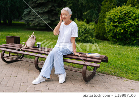 Unhappy elderly woman sitting in park bench feeling unwell, touching head, showing signs of pain and exhaustion, surrounded by nature. Reflects challenges of aging, mental health of senior healthcare 119997106