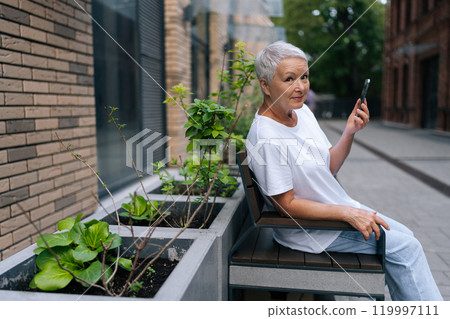 Portrait of elegant senior woman sitting on urban bench, holding smartphone and looking at camera with cute cheerful smile, enjoying free time in retirement outdoors. 119997111