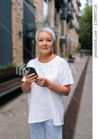 Vertical portrait of happy senior woman using typing smartphone while walking on street near modern building. Mature female tourist in white t-shirt reading writing text message, chatting online 119997116