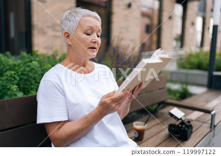 Side view of serene senior woman peacefully reading outdoors in city bench, embodying wisdom and experience. European gray-haired older female showcases happy retirement lifestyle. Side view of serene senior woman peacefully reading outdoors in city bench, embodying wisdom and experience. European gray-haired older female showcases happy retirement lifestyle. 119997122