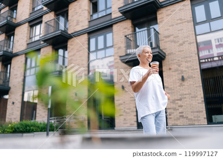 Low-angle view of confident European gray-haired older female strolling through urban setting, holding refreshing beverage and enjoying sights and sounds of city on summer day. Low-angle view of confident European gray-haired older female strolling through urban setting, holding refreshing beverage and enjoying sights and sounds of city on summer day. 119997127