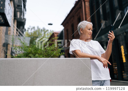 Relaxed gray-haired senior woman having video call, holding smartphone, gadget in hand, looking at screen, smiling, standing on background of modern European building on summer day. Relaxed gray-haired senior woman having video call, holding smartphone, gadget in hand, looking at screen, smiling, standing on background of modern European building on summer day. 119997128