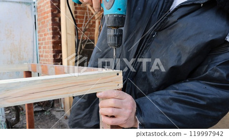 A man in dirty clothes using a screwdriver screws self-tapping screws into a wooden structure holding it with one hand on the street against a brick wall A man in dirty clothes using a screwdriver screws self-tapping screws into a wooden structure holding it with one hand on the street against a brick wall 119997942