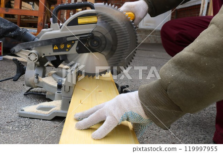 A worker wearing work clothes and construction gloves saws a light-colored wooden board with a hand-held circular saw A worker wearing work clothes and construction gloves saws a light-colored wooden board with a hand-held circular saw 119997955