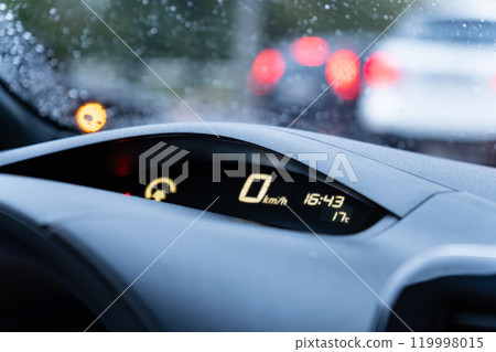 A car's dashboard features a speedometer reading zero while displaying the current time and temperature. Rain droplets are evident on the windshield, indicating wet weather 119998015