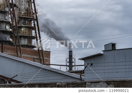 A factory emits thick smoke from its chimney, contrasting against a backdrop of overcast skies and nearby industrial structures, depicting a typical manufacturing environment 119998021