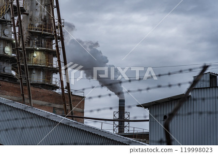 A large industrial facility releases smoke into the gray sky, showcasing the complexity of urban life amid towering structures and barbed wire A large industrial facility releases smoke into the gray sky, showcasing the complexity of urban life amid towering structures and barbed wire 119998023