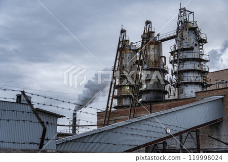 Tall smokestacks emit smoke into the gray sky, surrounded by industrial buildings. A conveyor belt runs through the foreground, enclosed by a barbed wire fence Tall smokestacks emit smoke into the gray sky, surrounded by industrial buildings. A conveyor belt runs through the foreground, enclosed by a barbed wire fence 119998024