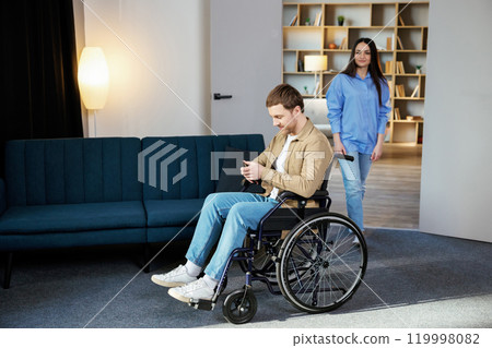 A cute young man in a wheelchair is holding a mobile phone while his girlfriend helps him move around the apartment 119998082