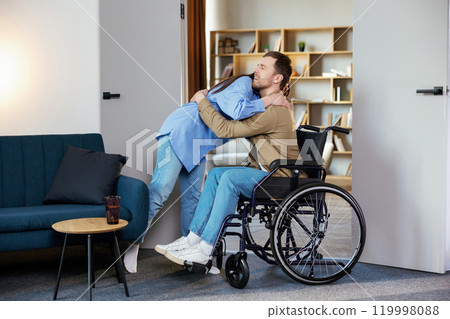 Young man in wheelchair and his wife hugging, standing in living room at home Young man in wheelchair and his wife hugging, standing in living room at home 119998088