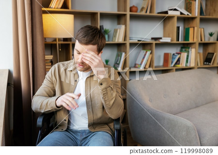 A young handsome man in a wheelchair looks out the window with a sad look A young handsome man in a wheelchair looks out the window with a sad look 119998089