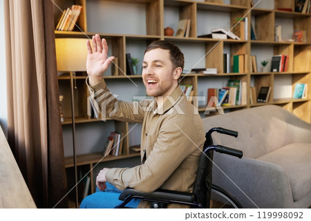 Man on wheelchair in room looking through window and smiling. Side view of young disabled patient in wheelchair looking out window 119998092