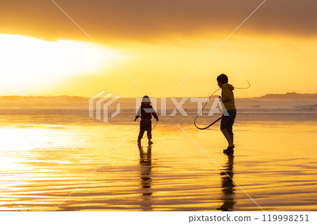 Silhouettes of two siblings playing with seaweeds on the beach in sunset. 119998251