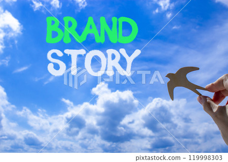 Brand story branding symbol. Concept words Brand story on beautiful blue sky. Beautiful blue sky clouds background. Hand holds wooden bird. Business brand story branding concept. Copy space. Brand story branding symbol. Concept words Brand story on beautiful blue sky. Beautiful blue sky clouds background. Hand holds wooden bird. Business brand story branding concept. Copy space. 119998303