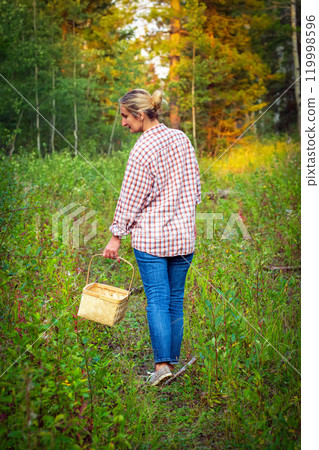 A woman in blue jeans and plaid shirt with a wicker basket is walking in the forest trail among trees and bushes and looking for mushrooms. 119998596