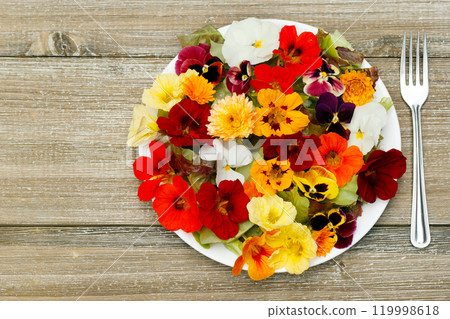 Beautiful healthy vegan salad of lettuce and colorful edible  flowers (nasturtium, calendula and pansy) on the wooden garden table with a fork on a side. 119998618
