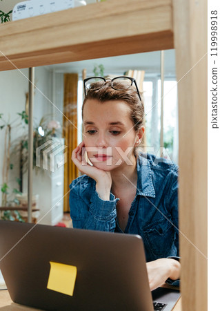European woman is sitting at desk in home office. looks at laptop screen monitoring finances. 119998918