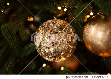 Close-up of a sparkling, beaded Christmas ornament on a green pine tree branch, with warm holiday lights in the background. 119999253
