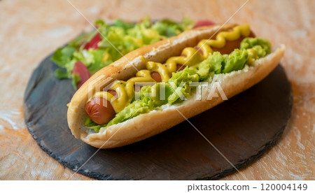 Hot dog on a bun, topped with mustard, guacamole and fresh salad. 120004149