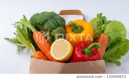 A paper bag filled with fresh vegetables including carrots bell peppers lettuce and a lemon is placed on a white surface against a white background 120004653