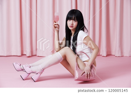 A Korean woman in her 20s with a champagne glass sitting on the pink floor wearing a Barbie Core look 120005145