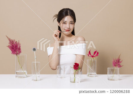 a woman in her 20s holding cosmetics in front of a table decorated with flowers 120006288