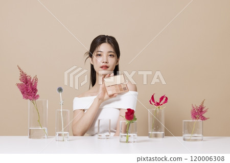 a woman in her 20s holding cosmetics in front of a table decorated with flowers 120006308