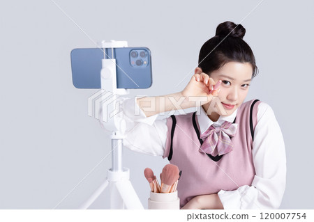 A teenage woman in school uniform laying out various cosmetics and filming a video with her smartphone 120007754