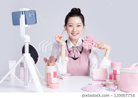 A teenage woman in school uniform laying out various cosmetics and filming a video with her smartphone 120007758