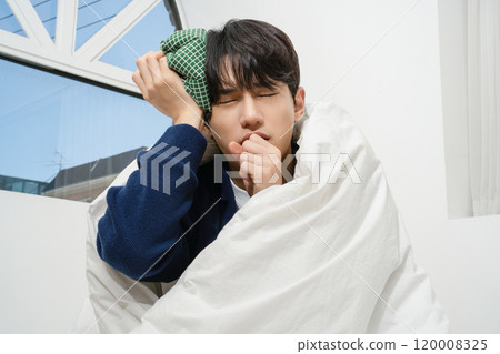 A person wrapped in a blanket and holding an ice pack on his forehead due to body aches A person wrapped in a blanket and holding an ice pack on his forehead due to body aches 120008325