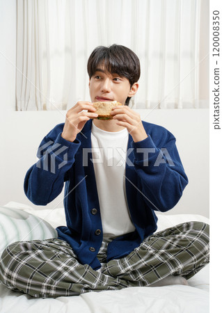 A man in his 20s sitting on the bed and eating bread 120008350