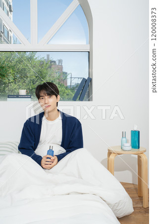 A man in his 20s sitting on a bed in front of a window and holding cosmetics 120008373