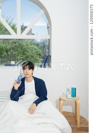 A man in his 20s sitting on a bed in front of a window and holding cosmetics 120008375