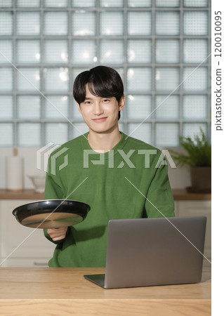A man in his 20s holding a frying pan and using a laptop 120010095