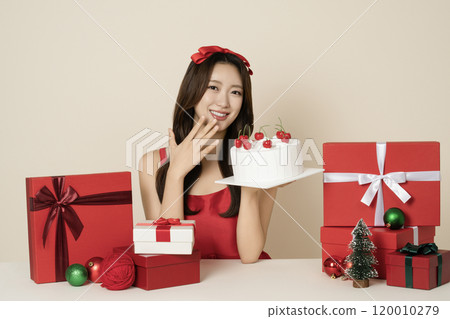 A woman in her 20s wearing a red dress posing with a cake surrounded by Christmas gifts and ornaments. 120010279