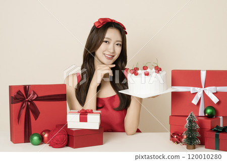 A woman in her 20s wearing a red dress posing with a cake surrounded by Christmas gifts and ornaments. 120010280