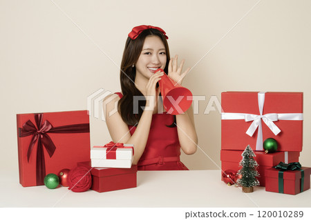 A woman in her 20s wearing a red dress holding a megaphone and surrounded by Christmas gifts and ornaments. 120010289