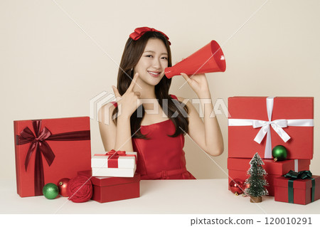 A woman in her 20s wearing a red dress holding a megaphone and surrounded by Christmas gifts and ornaments. A woman in her 20s wearing a red dress holding a megaphone and surrounded by Christmas gifts and ornaments. 120010291