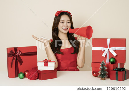 A woman in her 20s wearing a red dress holding a megaphone and surrounded by Christmas gifts and ornaments. A woman in her 20s wearing a red dress holding a megaphone and surrounded by Christmas gifts and ornaments. 120010292