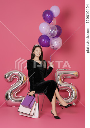 A woman in her 20s sitting and posing with a balloon and a shopping bag in front of a 25 number balloon. 120010404