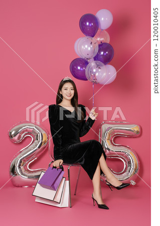 A woman in her 20s sitting and posing with a balloon and a shopping bag in front of a 25 number balloon. A woman in her 20s sitting and posing with a balloon and a shopping bag in front of a 25 number balloon. 120010405