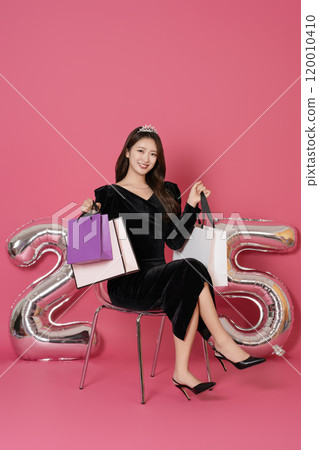 A woman in her 20s sitting and posing with a shopping bag in front of a 25 number balloon. 120010410