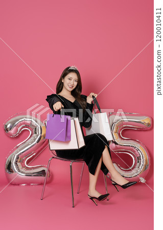 A woman in her 20s sitting and posing with a shopping bag in front of a 25 number balloon. 120010411