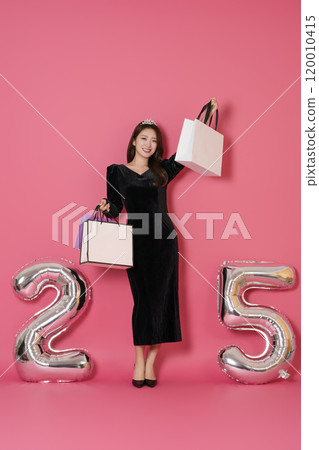 A woman in her 20s posing while holding a shopping bag in front of a 25 number balloon. 120010415
