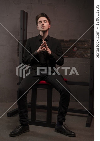 Confident young man in black suit sits in bdsm room 120011235