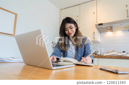 Young Freelance Woman Working from Home in Modern Kitchen with Laptop and Notebook, Smiling and Productive 120011565